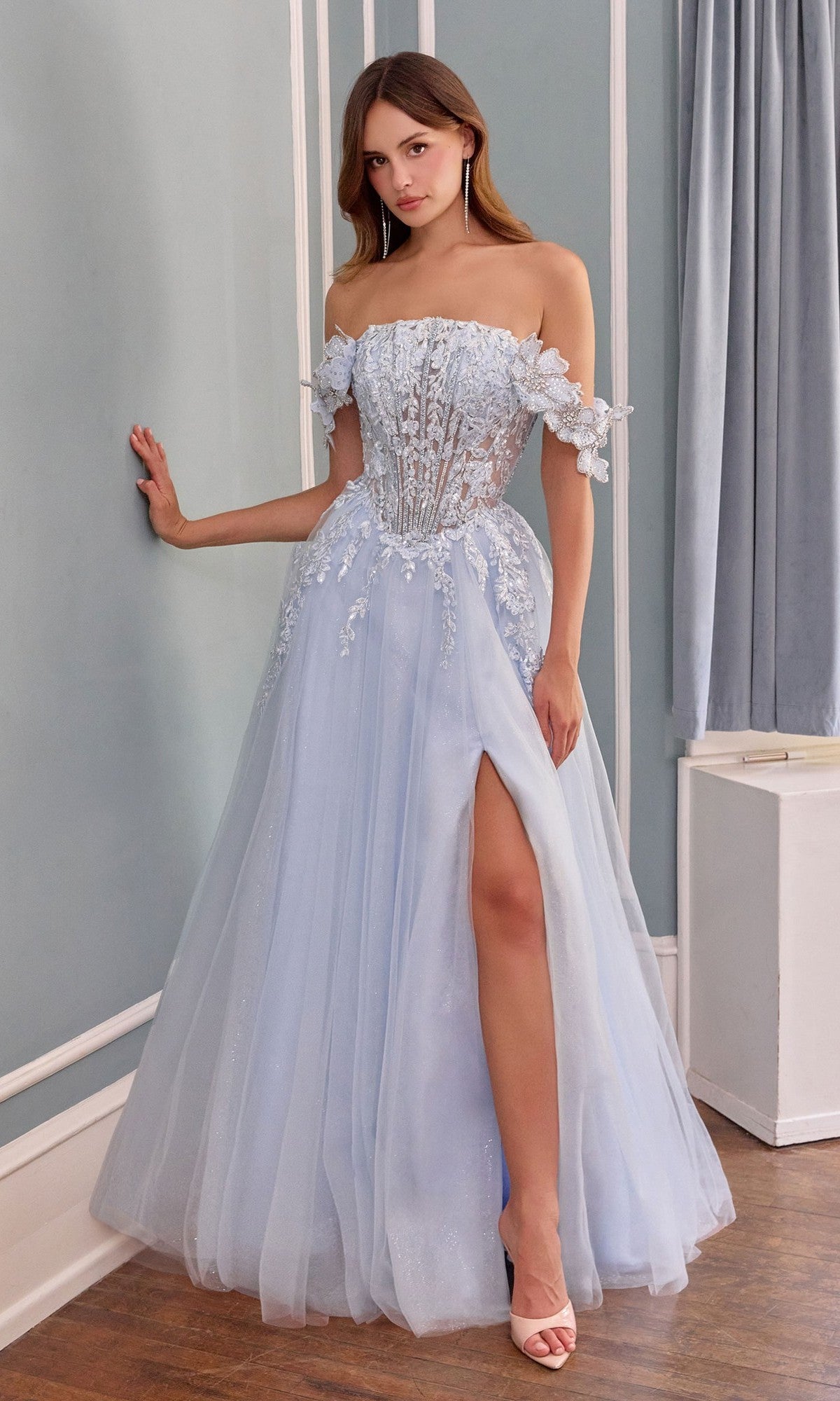 A woman wearing the Andrea and Leo A1512 long prom dress in light blue, featuring an off-shoulder a-line silhouette with 3D floral appliques, a sheer corset bodice, and high slit, stands indoors against a blue wall.